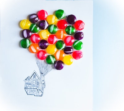 A playful arrangement of colorful candies placed strategically on a piece of paper, resembling balloons lifting a hand-drawn sketch of a house. The candies are vibrant and round, creating a whimsical and imaginative scene.