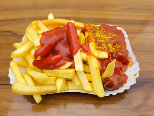 A serving of French fries topped with ketchup is placed beside pieces of sausage covered in curry powder and ketchup, all presented on a white paper plate on a wooden surface.