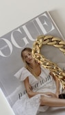 A magazine cover with the title 'Vogue' featuring a woman in a white lace dress with blonde hair. A large gold chain accessory is placed over part of the cover.