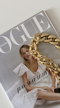A magazine cover with the title 'Vogue' featuring a woman in a white lace dress with blonde hair. A large gold chain accessory is placed over part of the cover.