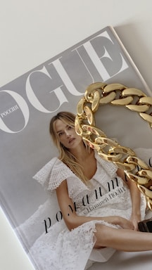 A magazine cover with the title 'Vogue' featuring a woman in a white lace dress with blonde hair. A large gold chain accessory is placed over part of the cover.