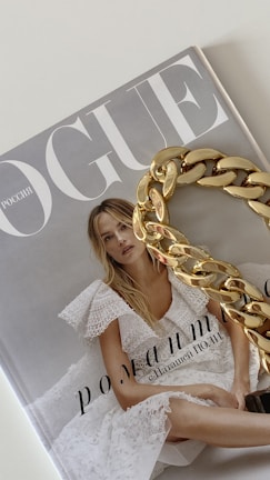 A magazine cover with the title 'Vogue' featuring a woman in a white lace dress with blonde hair. A large gold chain accessory is placed over part of the cover.