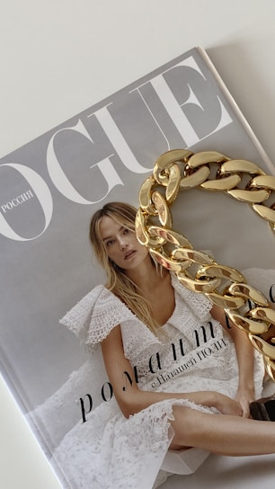 A magazine cover with the title 'Vogue' featuring a woman in a white lace dress with blonde hair. A large gold chain accessory is placed over part of the cover.