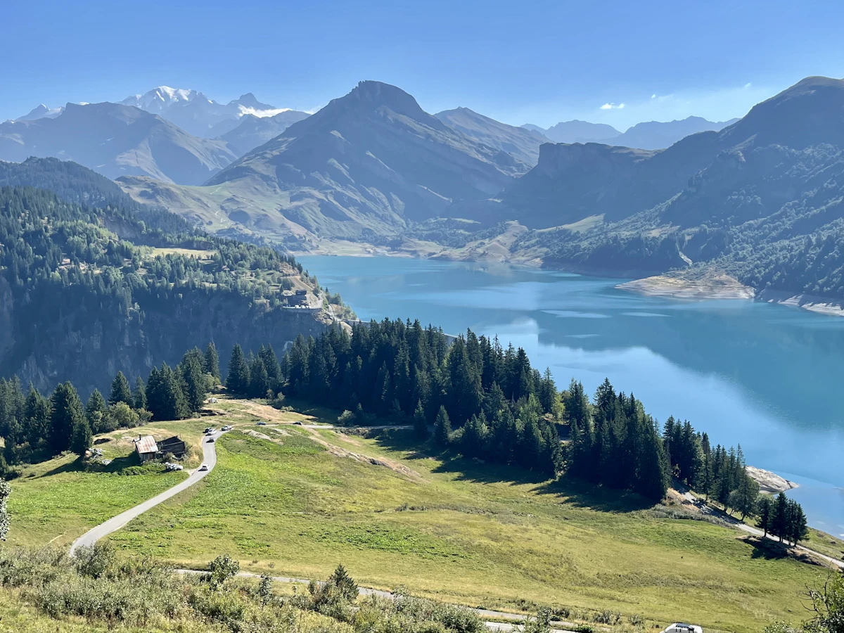 The lake below the Cormet de Roselend, under green mountains, with pines, a chalet and a track on the meadow above the water