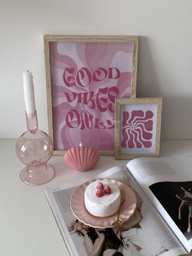 A decorative arrangement featuring two framed artworks with pink and white swirls and the text 'Good Vibes Only'. A single white candle in a pink glass candle holder and a pink seashell-shaped ornament are placed nearby. An open magazine showcases a page with fashion photography. On top of the magazine lies a small cake on a pink plate, adorned with raspberries and complemented by a gold fork.