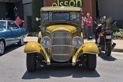A yellow vintage car with chrome detailing and round headlights is parked on a street. To its left, there is a blue classic car, and to its right, there is a motorcycle with a black and orange design. In the background, there is a wooden building with a green sign, and two women are standing near some potted plants. One of the women is wearing a pink top and a matching hat while the other is in casual clothing.