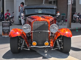 A vibrant, vintage car with a custom paint job featuring orange and red flames parked on a street. The car has a classic, retro design, with prominently displayed headlights and chrome detailing. There are several people casually seated in the background, some engaging in conversation, and a signboard to the left providing details about the car.