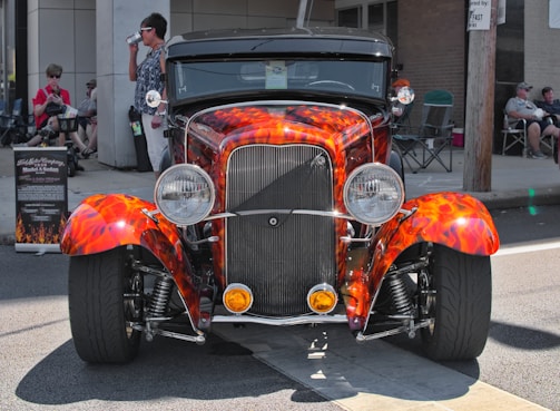 A vibrant, vintage car with a custom paint job featuring orange and red flames parked on a street. The car has a classic, retro design, with prominently displayed headlights and chrome detailing. There are several people casually seated in the background, some engaging in conversation, and a signboard to the left providing details about the car.