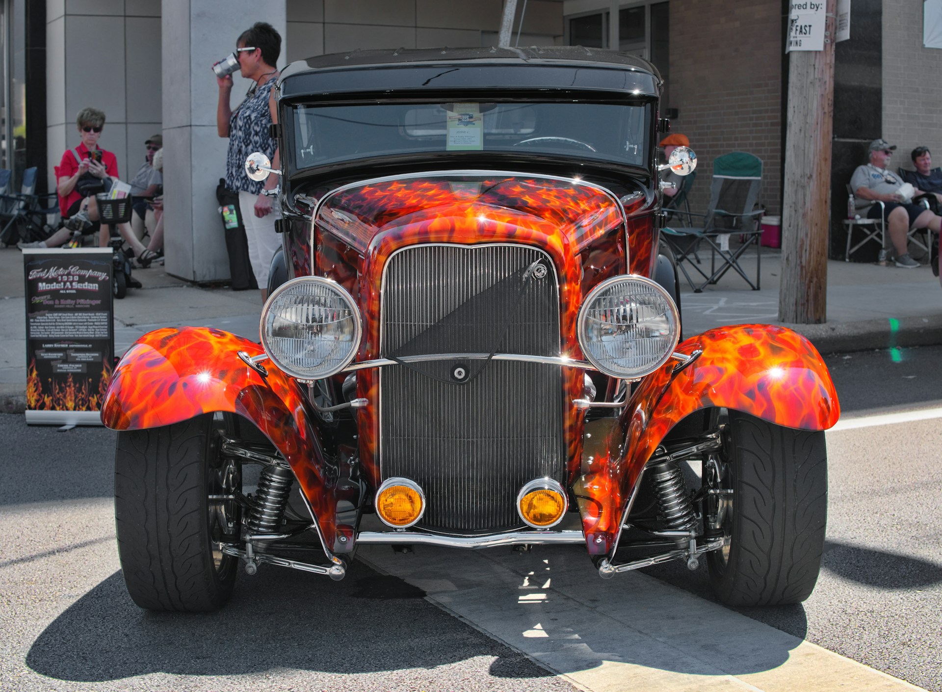 A gleaming vintage hot rod with polished chrome and vibrant flames painted along the sides, parked on a sunny street during a club meet.