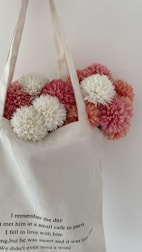 A white cloth bag with black text is filled with fluffy chrysanthemums. The flowers are in shades of white, pink, and light pink. The text on the bag reads: 'I remember the day I met him at a small cafe in Paris. I fell in love with him young, but he was sweet and it was true love. We didn't even need a word.' The background is plain and light-colored.