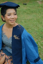 A person wearing a blue graduation gown and cap is sitting on grass. The gown has a logo that reads 'BINUS University.' The person is smiling and appears to be outdoors.