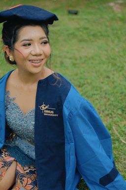 A person wearing a blue graduation gown and cap is sitting on grass. The gown has a logo that reads 'BINUS University.' The person is smiling and appears to be outdoors.