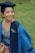 A person wearing a blue graduation gown and cap is sitting on grass. The gown has a logo that reads 'BINUS University.' The person is smiling and appears to be outdoors.