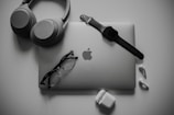 A neatly organized collection of tech gadgets featuring a closed laptop with the Apple logo, over-ear headphones, a smartwatch with a black band, a pair of black-framed glasses, and white wireless earbuds along with their charging case.