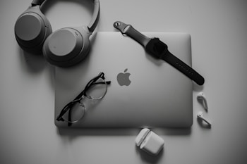 A neatly organized collection of tech gadgets featuring a closed laptop with the Apple logo, over-ear headphones, a smartwatch with a black band, a pair of black-framed glasses, and white wireless earbuds along with their charging case.