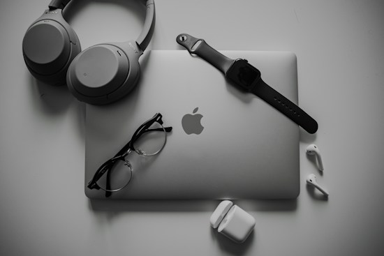 A neatly organized collection of tech gadgets featuring a closed laptop with the Apple logo, over-ear headphones, a smartwatch with a black band, a pair of black-framed glasses, and white wireless earbuds along with their charging case.