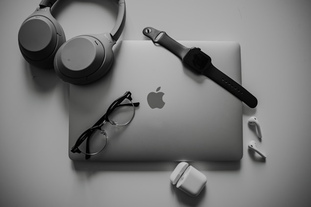 A neatly organized collection of tech gadgets featuring a closed laptop with the Apple logo, over-ear headphones, a smartwatch with a black band, a pair of black-framed glasses, and white wireless earbuds along with their charging case.