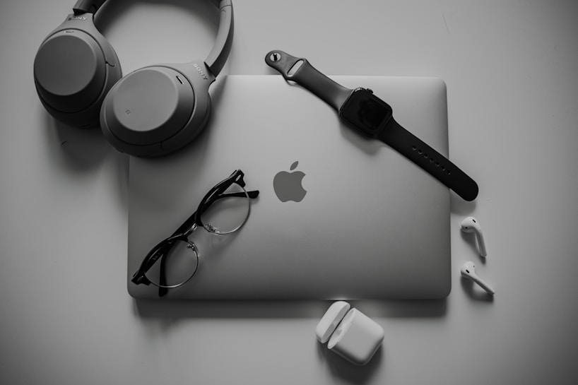 A neatly organized collection of tech gadgets featuring a closed laptop with the Apple logo, over-ear headphones, a smartwatch with a black band, a pair of black-framed glasses, and white wireless earbuds along with their charging case.