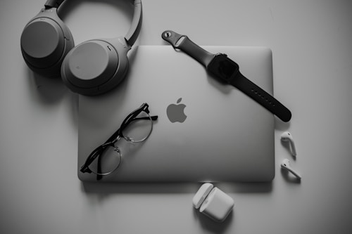 A neatly organized collection of tech gadgets featuring a closed laptop with the Apple logo, over-ear headphones, a smartwatch with a black band, a pair of black-framed glasses, and white wireless earbuds along with their charging case.