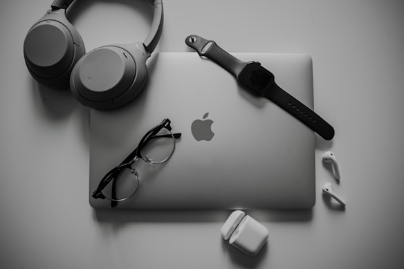A neatly organized collection of tech gadgets featuring a closed laptop with the Apple logo, over-ear headphones, a smartwatch with a black band, a pair of black-framed glasses, and white wireless earbuds along with their charging case.