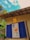 A vibrant mural is displayed between two blue shutters on a rustic building with a weathered roof. The mural features abstract floral elements with bright colors, set against a sky with fluffy clouds. A small hanging lantern adds warmth to the scene, and greenery surrounds the area.