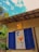 A vibrant mural is displayed between two blue shutters on a rustic building with a weathered roof. The mural features abstract floral elements with bright colors, set against a sky with fluffy clouds. A small hanging lantern adds warmth to the scene, and greenery surrounds the area.