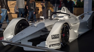 A sleek white race car with aerodynamic design, prominently displaying brand logos like Porsche, Boss, and Mobil, is showcased on a platform. Several people stand around the car, wearing casual attire, suggesting an event or exhibition setting.
