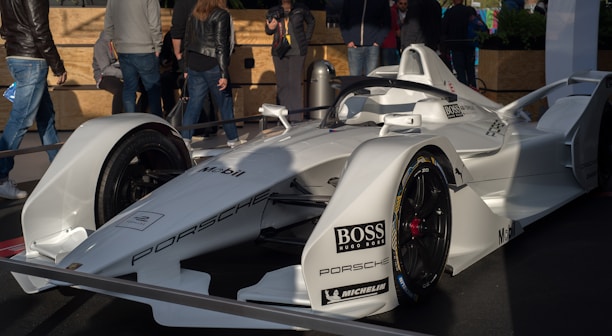 A sleek white race car with aerodynamic design, prominently displaying brand logos like Porsche, Boss, and Mobil, is showcased on a platform. Several people stand around the car, wearing casual attire, suggesting an event or exhibition setting.