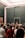 A large group of people gathered in an art gallery, many of them taking photos with their phones. Several paintings, including a famous portrait, are visible on the walls. The space has a modern and minimalistic design with dark walls and a grid-patterned glass ceiling.