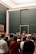A large group of people gathered in an art gallery, many of them taking photos with their phones. Several paintings, including a famous portrait, are visible on the walls. The space has a modern and minimalistic design with dark walls and a grid-patterned glass ceiling.