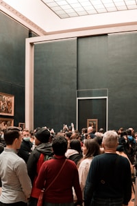 A large group of people gathered in an art gallery, many of them taking photos with their phones. Several paintings, including a famous portrait, are visible on the walls. The space has a modern and minimalistic design with dark walls and a grid-patterned glass ceiling.