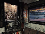 A room filled with Eastern art and sculptures, featuring a prominent bronze statue of a meditative figure, possibly Buddha, surrounded by smaller similar statues and various decorative items. A colorful tapestry depicting a deity is hung near a framed panoramic painting of a serene lake with a solitary boat, set against a backdrop of jagged mountains under a dramatic sky.