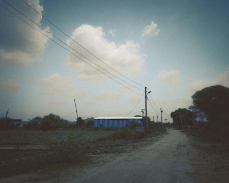 A rural scene with a dirt road leading towards a distant warehouse structure surrounded by greenery. Power lines stretch across the scene, under a partially cloudy sky.