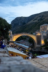 A historic stone bridge stretches across a river, illuminated by warm lights as dusk falls. People are gathered on and around the bridge, indicating a popular tourist or cultural spot. Surrounding the scene are rustic buildings with sloped roofs and a backdrop of large rocky hills.