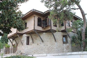 A traditional stone house with wooden upper floors and open windows is surrounded by lush green trees. The building features rustic wooden beams and a small balcony.
