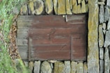 A rustic wooden door with visible grain and aged texture, set into a stone wall composed of unevenly stacked rocks. The stones are covered with patches of moss and lichen, adding earthy tones to the scene. Surrounding leaves and greenery frame the wooden door, suggesting it is part of an outdoor environment.