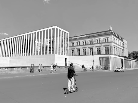 A person rides an electric scooter on a wide street in an urban area, towards modern and classical style buildings. Several pedestrians are walking on the sidewalk, and a car is parked near the building. The sky is clear, indicating sunny weather.
