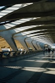 The architecture features a spacious, modern walkway with large, curved concrete beams and a roof allowing natural light to cast dramatic shadows on the floor. A few people are walking along the path, contributing to the sense of movement and activity.