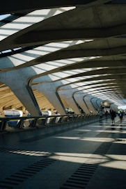 The architecture features a spacious, modern walkway with large, curved concrete beams and a roof allowing natural light to cast dramatic shadows on the floor. A few people are walking along the path, contributing to the sense of movement and activity.