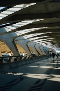 The architecture features a spacious, modern walkway with large, curved concrete beams and a roof allowing natural light to cast dramatic shadows on the floor. A few people are walking along the path, contributing to the sense of movement and activity.
