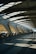 The architecture features a spacious, modern walkway with large, curved concrete beams and a roof allowing natural light to cast dramatic shadows on the floor. A few people are walking along the path, contributing to the sense of movement and activity.