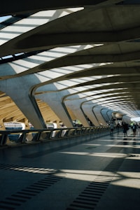 The architecture features a spacious, modern walkway with large, curved concrete beams and a roof allowing natural light to cast dramatic shadows on the floor. A few people are walking along the path, contributing to the sense of movement and activity.