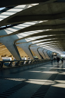 The architecture features a spacious, modern walkway with large, curved concrete beams and a roof allowing natural light to cast dramatic shadows on the floor. A few people are walking along the path, contributing to the sense of movement and activity.