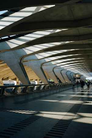 The architecture features a spacious, modern walkway with large, curved concrete beams and a roof allowing natural light to cast dramatic shadows on the floor. A few people are walking along the path, contributing to the sense of movement and activity.