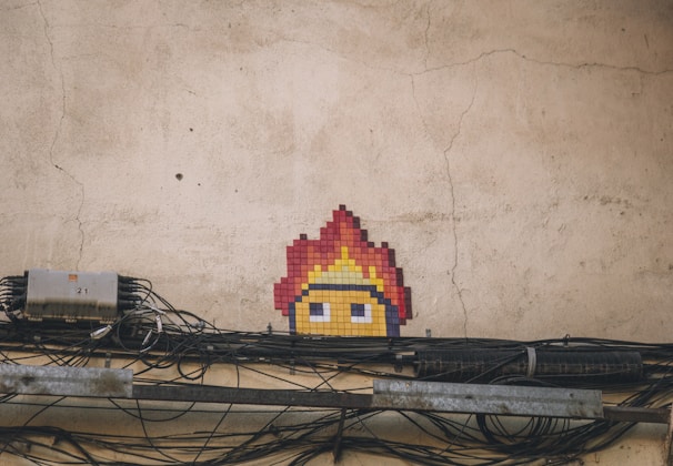 A mosaic art piece of a pixelated character's head with fiery hair on an old, cracked wall. Below are multiple tangled and bundled black cables running horizontally along the wall with an electrical box labeled '21' to the left.