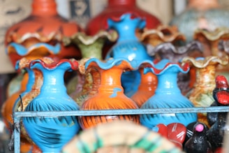 Brightly colored ceramic vases with wavy edges are scattered among a variety of vibrant pottery items. The vases feature shades of blue and orange, complemented by other decorative pottery pieces with red and green hues.