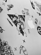 A collection of black and white stickers on a flat surface, possibly a laptop. The central sticker features an astronaut holding a light or spark, with a background of stars and a triangular border. Surrounding the astronaut are various other stickers including flowers, a snake, a bullet, a syringe, an hourglass, a bag labeled 'Karma', a tombstone with the number '666', and the Apple logo.