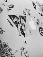 A collection of black and white stickers on a flat surface, possibly a laptop. The central sticker features an astronaut holding a light or spark, with a background of stars and a triangular border. Surrounding the astronaut are various other stickers including flowers, a snake, a bullet, a syringe, an hourglass, a bag labeled 'Karma', a tombstone with the number '666', and the Apple logo.