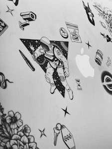 A collection of black and white stickers on a flat surface, possibly a laptop. The central sticker features an astronaut holding a light or spark, with a background of stars and a triangular border. Surrounding the astronaut are various other stickers including flowers, a snake, a bullet, a syringe, an hourglass, a bag labeled 'Karma', a tombstone with the number '666', and the Apple logo.