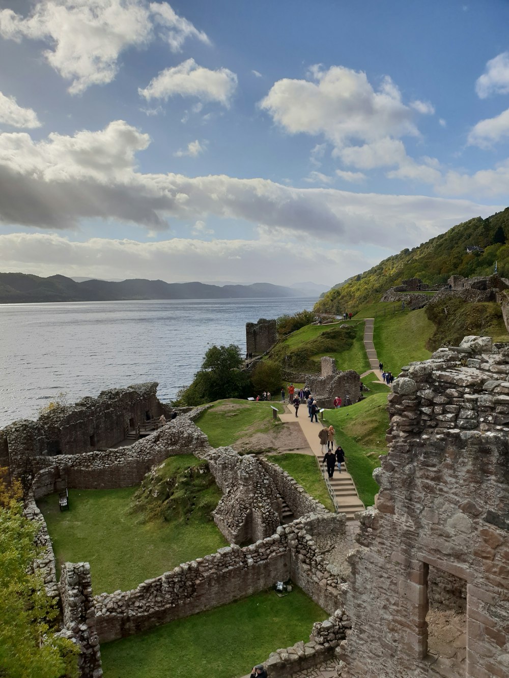 a stone wall with a walkway and people walking on it by a body of water with Urquhart Castle in the background